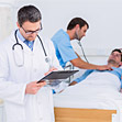 immagine con paziente a letto e due medici: uno lo visita, l'altro scrive in cartella clinica